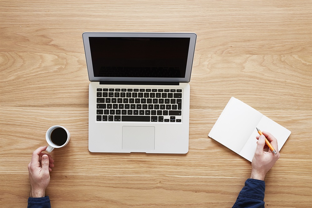 Foto. Tagen uppifrån. En laptop står på ett bord. I nederkant syns två händer, en håller i en kaffekopp och en skriver i en anteckningsbok.